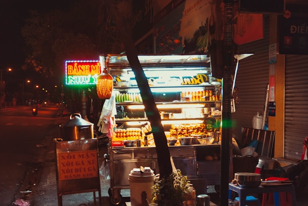 A brightly lit street food stall at night displays an assortment of snacks and beverages. Neon signs, including one with the words 'Bánh Mì', contribute to the vibrant atmosphere. A large pot and other cooking items are visible around the stall, which is situated on a quiet street with minimal traffic.