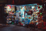 A small, bustling street shop illuminated by bright lights offers a wide variety of snacks, drinks, and other goods. The shop is filled with colorful packaging, with a refrigerator visible to the right and various merchandise hanging or stacked around. A person is seated on a stool in front of the shop, with plastic chairs scattered nearby.