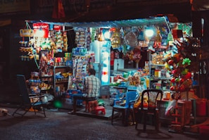A small, bustling street shop illuminated by bright lights offers a wide variety of snacks, drinks, and other goods. The shop is filled with colorful packaging, with a refrigerator visible to the right and various merchandise hanging or stacked around. A person is seated on a stool in front of the shop, with plastic chairs scattered nearby.