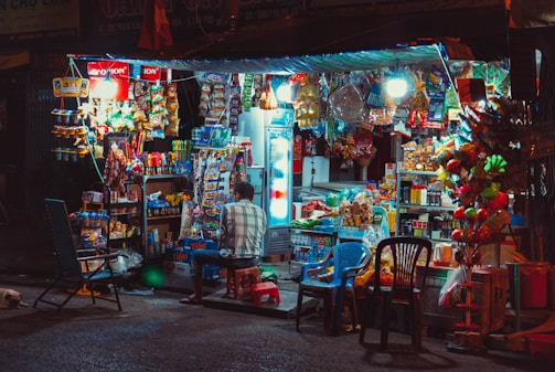 A small, bustling street shop illuminated by bright lights offers a wide variety of snacks, drinks, and other goods. The shop is filled with colorful packaging, with a refrigerator visible to the right and various merchandise hanging or stacked around. A person is seated on a stool in front of the shop, with plastic chairs scattered nearby.