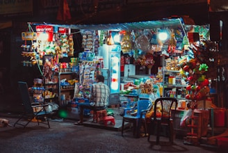 A small, bustling street shop illuminated by bright lights offers a wide variety of snacks, drinks, and other goods. The shop is filled with colorful packaging, with a refrigerator visible to the right and various merchandise hanging or stacked around. A person is seated on a stool in front of the shop, with plastic chairs scattered nearby.