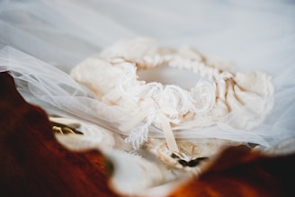 A delicate lace garter intertwined with tulle fabric lies atop a wooden surface, featuring intricate embroidery and a small ribbon bow.