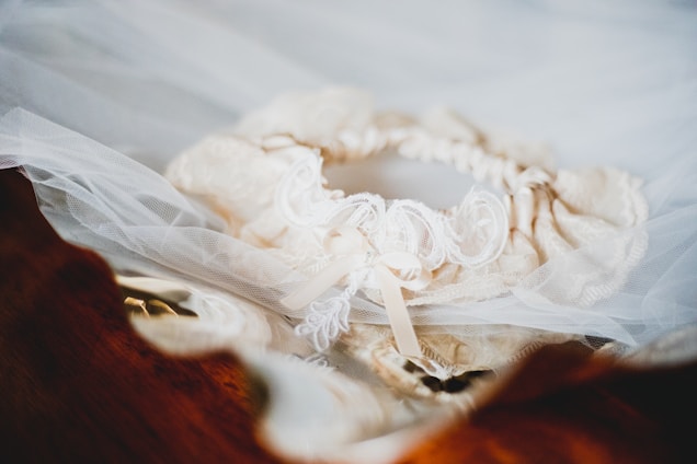 A delicate lace garter intertwined with tulle fabric lies atop a wooden surface, featuring intricate embroidery and a small ribbon bow.