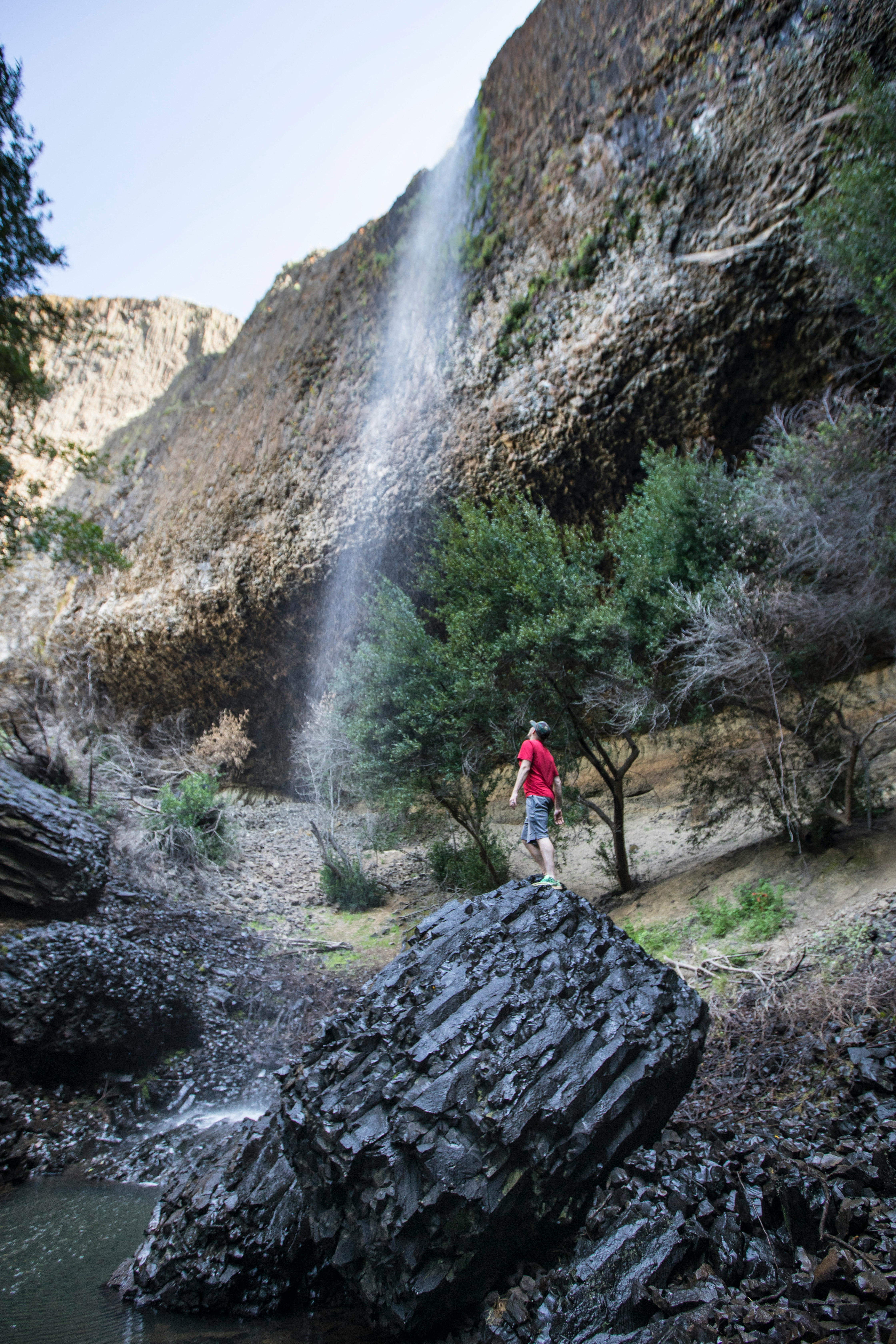 man in red t-shirt standing on rock near waterfalls