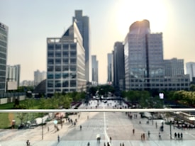 A cityscape featuring tall modern skyscrapers with glass facades under a bright sky, where sunlight is visible near the top of the tallest building. Below, a busy plaza with people walking creates a lively urban atmosphere. Trees line the pathways, adding a touch of greenery to the scene.