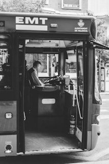 A black and white photograph captures the interior of a bus with a driver seated at the steering wheel. The bus is labeled with 'EMT' and 'Ajuntament de Valencia' on the front, indicating a public transport service in Valencia. The image shows details like the driver's seat, the dashboard controls, and a fire extinguisher near the entrance.