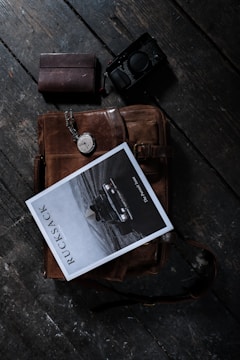 A leather bag lies on a dark wooden surface alongside a closed leather notebook, a vintage pocket watch, a magazine titled 'Rucksack', and a camera. The lighting creates a moody, vintage aesthetic.