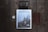 A flat lay photograph featuring a magazine titled 'Rucksack' with a cover image of a sailing ship navigating through icy waters near towering mountains. Positioned on a dark wooden surface, a vintage pocket watch with an attached chain is seen to the left, while a black camera is placed above. To the right, a closed brown leather notebook rests parallel to the magazine.