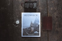 A flat lay photograph featuring a magazine titled 'Rucksack' with a cover image of a sailing ship navigating through icy waters near towering mountains. Positioned on a dark wooden surface, a vintage pocket watch with an attached chain is seen to the left, while a black camera is placed above. To the right, a closed brown leather notebook rests parallel to the magazine.