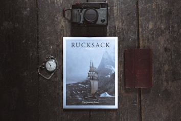A flat lay photograph featuring a magazine titled 'Rucksack' with a cover image of a sailing ship navigating through icy waters near towering mountains. Positioned on a dark wooden surface, a vintage pocket watch with an attached chain is seen to the left, while a black camera is placed above. To the right, a closed brown leather notebook rests parallel to the magazine.