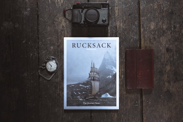 A flat lay photograph featuring a magazine titled 'Rucksack' with a cover image of a sailing ship navigating through icy waters near towering mountains. Positioned on a dark wooden surface, a vintage pocket watch with an attached chain is seen to the left, while a black camera is placed above. To the right, a closed brown leather notebook rests parallel to the magazine.