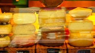 Various types of cheeses are stacked and arranged on wicker baskets, with labels indicating their names. The cheeses have different shapes and textures, and a warm, yellow and orange lighting enhances the presentation.