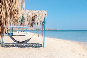 A serene beach scene with a hammock strung between two wooden posts under a thatched roof. The sand is light and clean, leading to a calm, blue ocean under a clear, sunny sky. There are multiple similar structures visible in the distance along the shoreline.
