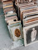 A collection of vintage framed photographs is arranged on the ground. The frames vary in style and size, showcasing sepia-toned portraits, including a child in traditional attire and a person in formal clothing. The setting appears to be an outdoor market or antique shop.