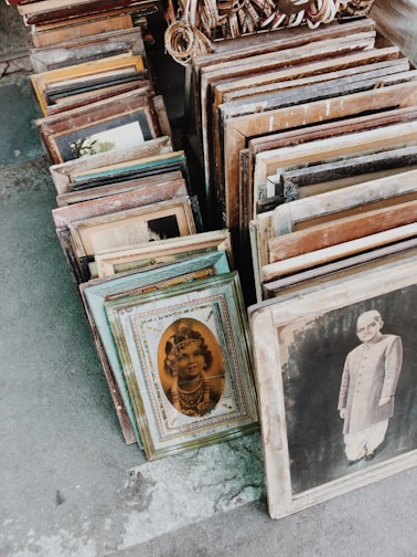 A collection of vintage framed photographs is arranged on the ground. The frames vary in style and size, showcasing sepia-toned portraits, including a child in traditional attire and a person in formal clothing. The setting appears to be an outdoor market or antique shop.