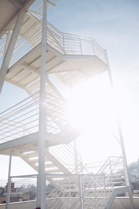 A metal staircase with multiple flights and railings, partially obscured by the bright sunlight. The structure appears to be outdoors with a clear blue sky in the background and some buildings visible at the bottom.