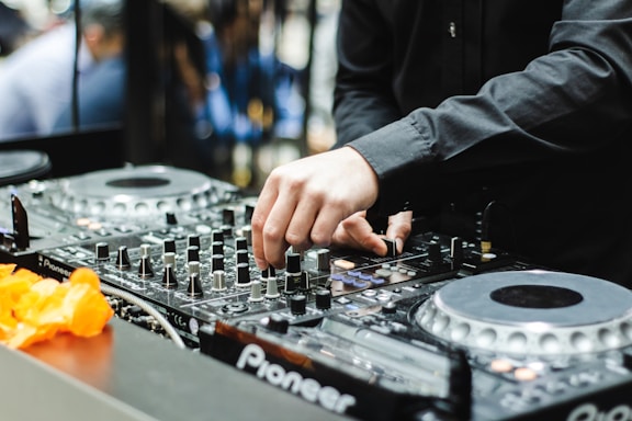 A DJ is adjusting the controls on a mixer, surrounded by professional audio equipment. The scene is dynamic, suggesting an active performance or sound mixing session.