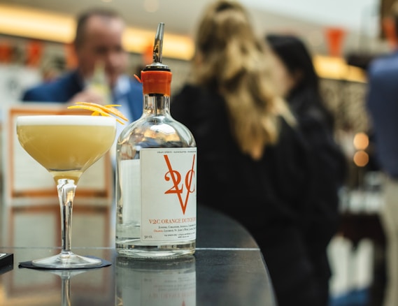 A bottle of V2C Orange Dutch Dry Gin and a cocktail glass with a frothy drink topped with a twist of orange peel are placed on a sleek, reflective table surface. In the background, several people are blurred, suggesting a social gathering or event setting.