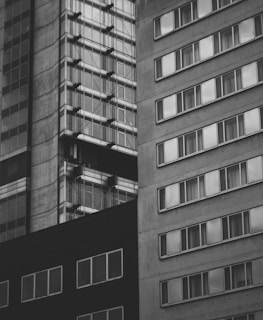 A black and white image featuring modern high-rise buildings with rectangular windows. The structures are closely packed together, showcasing a mix of concrete and glass materials, emphasizing urban architecture.