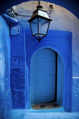 A vibrant blue arched doorway with intricate patterns around the door area. A classic black lantern hangs above, casting subtle shadows. The walls and surroundings are also painted in rich blue tones, creating a harmonious and calming visual experience.