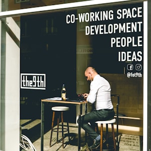 A person is sitting at an outdoor table in a co-working space, using a laptop. They appear focused as they work, with headphones on. The space has large windows with visible signage advertising development, people, and ideas, as well as social media symbols.