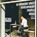 A person is sitting at an outdoor table in a co-working space, using a laptop. They appear focused as they work, with headphones on. The space has large windows with visible signage advertising development, people, and ideas, as well as social media symbols.