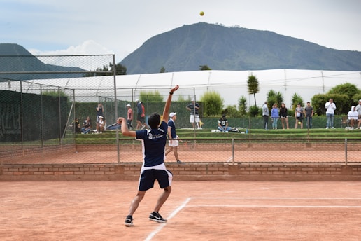 A tennis player is captured in mid-serve on a clay court, surrounded by a chain-link fence. Spectators are visible in the background, alongside green foliage and a white structure, likely a part of a sports facility, with a mountain under a cloudy sky in the distance.