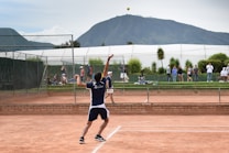 A tennis player is captured in mid-serve on a clay court, surrounded by a chain-link fence. Spectators are visible in the background, alongside green foliage and a white structure, likely a part of a sports facility, with a mountain under a cloudy sky in the distance.