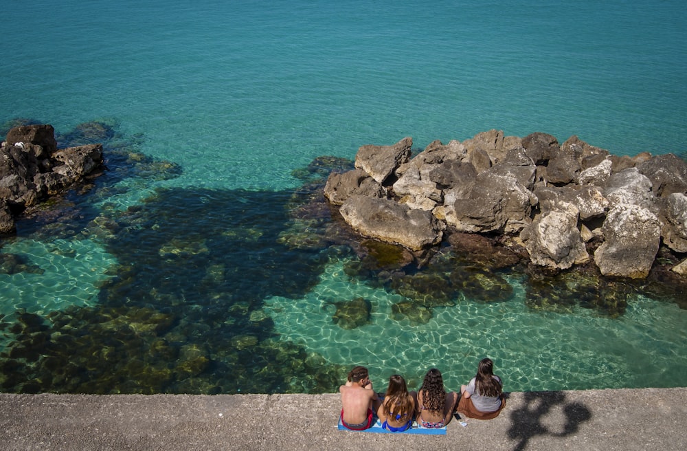 aerial photography of four people sitting on concrete pavement in front of seashore during daytime