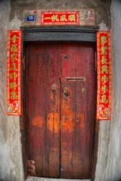 A wooden door with a rustic appearance, featuring traditional red Chinese calligraphy banners on each side and above the door, which are often associated with good luck and prosperity. The number 15 is displayed on a small blue plaque beside the top banner.