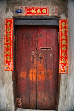 A wooden door with a rustic appearance, featuring traditional red Chinese calligraphy banners on each side and above the door, which are often associated with good luck and prosperity. The number 15 is displayed on a small blue plaque beside the top banner.