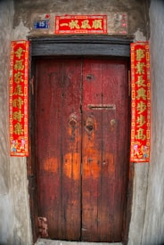 A wooden door with a rustic appearance, featuring traditional red Chinese calligraphy banners on each side and above the door, which are often associated with good luck and prosperity. The number 15 is displayed on a small blue plaque beside the top banner.