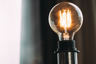 A close-up view of an illuminated incandescent light bulb with a visible filament. The bulb is mounted on a metallic base, and a soft, diffused background with vertical stripes adds a subtle contrast.