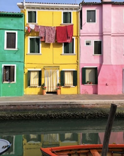 Brightly colored buildings line a waterfront with green, yellow, and pink facades. Clothes hang on a line across the yellow building. There are potted plants in front of one door, and a canal with a boat in the foreground.