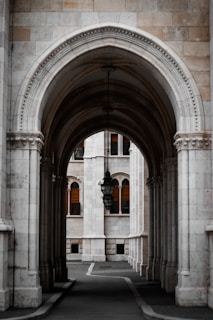 A beautifully ornate stone archway leads to a courtyard with tall columns and a classic lantern hanging in the center. The architecture features intricate detailing and symmetry, with the building's facade showcasing arched windows accented by wooden shutters.