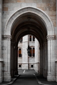 A beautifully ornate stone archway leads to a courtyard with tall columns and a classic lantern hanging in the center. The architecture features intricate detailing and symmetry, with the building's facade showcasing arched windows accented by wooden shutters.