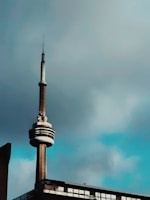 A tall communication tower with a distinctive circular structure near the top stands against a cloudy sky. The lower part of the tower is connected to a building with large windows.