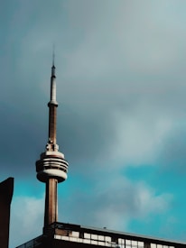 A tall communication tower with a distinctive circular structure near the top stands against a cloudy sky. The lower part of the tower is connected to a building with large windows.