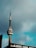 A tall communication tower with a distinctive circular structure near the top stands against a cloudy sky. The lower part of the tower is connected to a building with large windows.