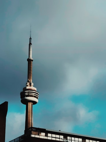 A tall communication tower with a distinctive circular structure near the top stands against a cloudy sky. The lower part of the tower is connected to a building with large windows.