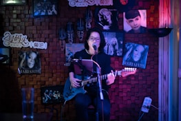 A person is playing an electric guitar and singing into a microphone in a cozy, dimly lit room decorated with music posters, including some of famous artists. The backdrop includes wooden paneling and colorful lighting.