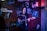 A person is playing an electric guitar and singing into a microphone in a cozy, dimly lit room decorated with music posters, including some of famous artists. The backdrop includes wooden paneling and colorful lighting.