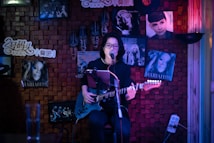 A person is playing an electric guitar and singing into a microphone in a cozy, dimly lit room decorated with music posters, including some of famous artists. The backdrop includes wooden paneling and colorful lighting.