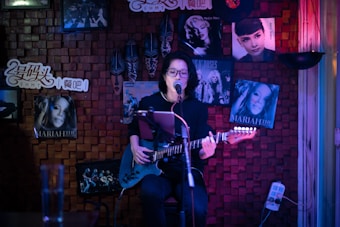 A person is playing an electric guitar and singing into a microphone in a cozy, dimly lit room decorated with music posters, including some of famous artists. The backdrop includes wooden paneling and colorful lighting.