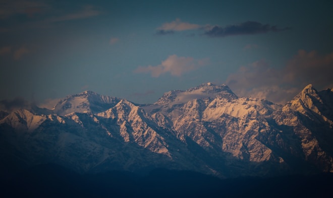 Snow-covered mountains glow under the warm hues of a sunset. Wispy clouds float above, casting soft shadows on the rugged peaks which are accentuated by the fading light.