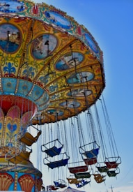 A colorful amusement park ride with swinging chairs hanging from a decorated canopy. The canopy features intricate designs and paintings of scenic views. The chairs are suspended on long chains and are painted in vibrant colors like blue, red, yellow, and green.