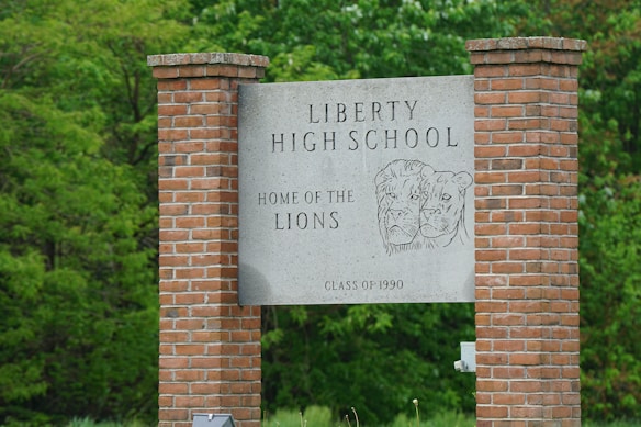 A large rectangular stone sign is held up by two red brick pillars. The text on the sign reads 'Liberty High School Home of the Lions' with a drawing of two lion heads. Below, it states 'Class of 1990'. The sign is set against a background of lush green trees, suggesting a school setting.