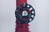 A close-up of a dark green valve wheel, mounted on a red pipe. The wheel has a central label with the word 'HYDRO'. The pipe has a textured surface with multiple facets and appears to be part of a larger industrial system.