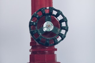 A close-up of a dark green valve wheel, mounted on a red pipe. The wheel has a central label with the word 'HYDRO'. The pipe has a textured surface with multiple facets and appears to be part of a larger industrial system.