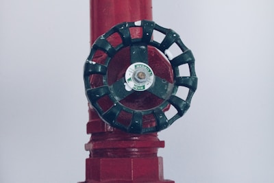 A close-up of a dark green valve wheel, mounted on a red pipe. The wheel has a central label with the word 'HYDRO'. The pipe has a textured surface with multiple facets and appears to be part of a larger industrial system.